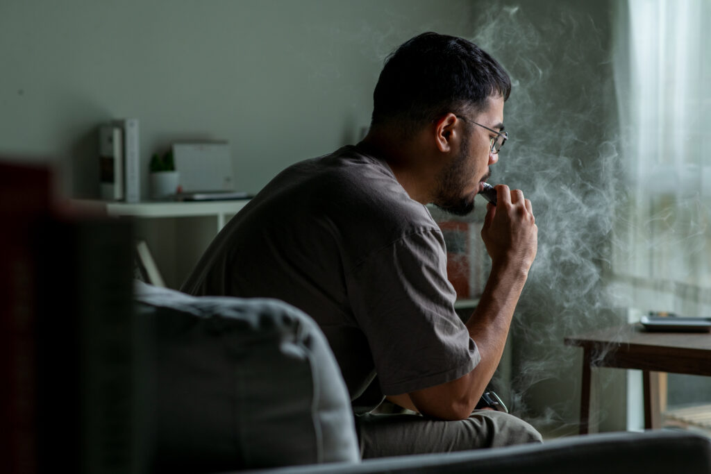 Young Asian man sitting on a sofa at home, Vaping, looking sad and exhausted, struggling with depression and mental health issues.
